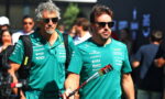 Two men in teal team shirts with sponsor logos walk side by side at an event, wearing sunglasses and carrying bags and a lanyard/flag pole.