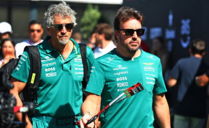Two men in teal team shirts with sponsor logos walk side by side at an event, wearing sunglasses and carrying bags and a lanyard/flag pole.
