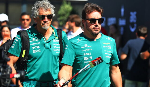 Two men in teal team shirts with sponsor logos walk side by side at an event, wearing sunglasses and carrying bags and a lanyard/flag pole.