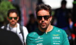 Man in a green racing team shirt with sponsor logos and sunglasses, outdoors in a crowd.