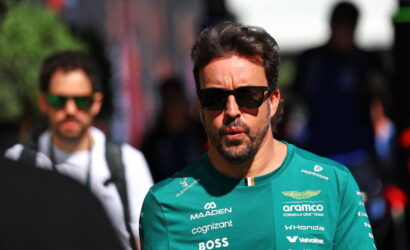 Man in a green racing team shirt with sponsor logos and sunglasses, outdoors in a crowd.