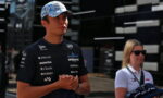 Young racing driver in a sponsor logos jersey and blue cap standing with hands clasped at a circuits paddock