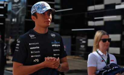 Young racing driver in a sponsor logos jersey and blue cap standing with hands clasped at a circuits paddock