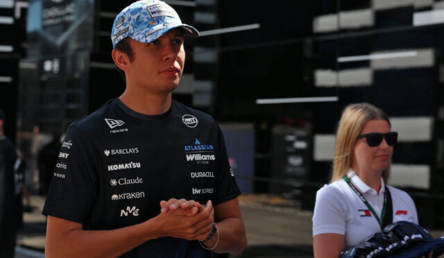 Young racing driver in a sponsor logos jersey and blue cap standing with hands clasped at a circuits paddock