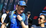 Male race car driver in a blue sponsor shirt and cap smiles while adjusting his backpack at a motorsport event.