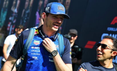 Male race car driver in a blue sponsor shirt and cap smiles while adjusting his backpack at a motorsport event.