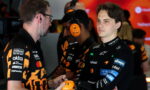 Two team members in black-and-orange uniforms talk in a garage as one hands a bright orange headset to a young driver in a black racing suit with sponsor logos; red screens in the background.
