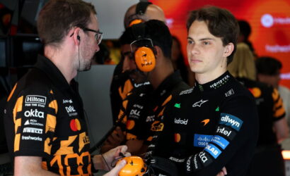 Two team members in black-and-orange uniforms talk in a garage as one hands a bright orange headset to a young driver in a black racing suit with sponsor logos; red screens in the background.