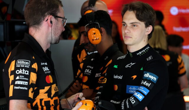 Two team members in black-and-orange uniforms talk in a garage as one hands a bright orange headset to a young driver in a black racing suit with sponsor logos; red screens in the background.