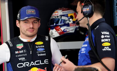 Oracle Red Bull Racing crew member with cap speaks with teammate in headset; helmet visible in background.
