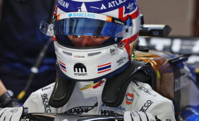 Close-up of a race car driver in a white helmet and suit, gripping the steering wheel in a Formula 1 cockpit with sponsor logos visible on the helmet.