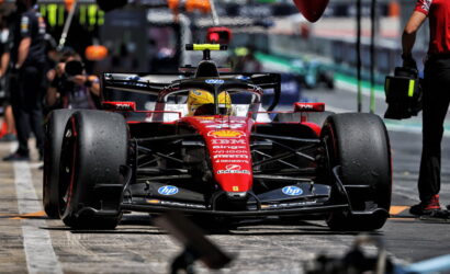 Red Ferrari Formula 1 car on the pit lane ready for a stop, driver in yellow helmet visible.