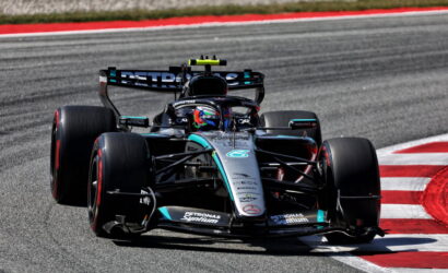 Mercedes-AMG Petronas Formula 1 car navigating a red-and-white curbed corner on a racetrack, driver in helmet visible behind the cockpit.