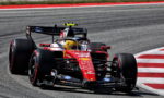 Red Ferrari Formula 1 car taking a sharp corner on a race track, front wheels turned and driver wearing a yellow helmet visible in the cockpit.