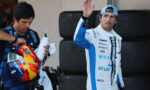 Racing drivers in a pit area: one in a white-and-blue suit waves, while the teammate holds a colorful helmet and a water bottle nearby of stacked tires in the background.