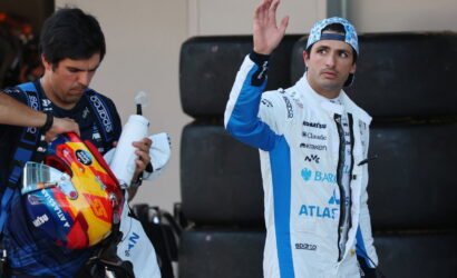 Racing drivers in a pit area: one in a white-and-blue suit waves, while the teammate holds a colorful helmet and a water bottle nearby of stacked tires in the background.