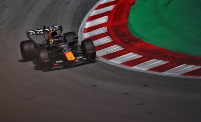 Red Bull Formula 1 car taking a sharp corner on a race track, driver in helmet visible, red-and-white striped curbs nearby