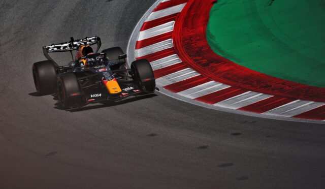 Red Bull Formula 1 car taking a sharp corner on a race track, driver in helmet visible, red-and-white striped curbs nearby