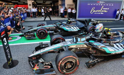 Mercedes Formula 1 cars lined up in the pit lane with a cheering crowd taking photos, a large Barcelona banner in the background.