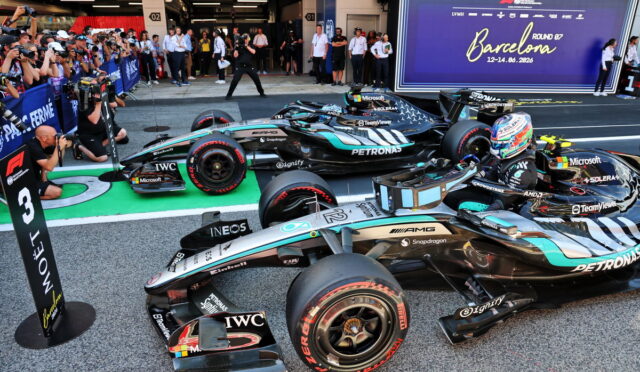 Mercedes Formula 1 cars lined up in the pit lane with a cheering crowd taking photos, a large Barcelona banner in the background.