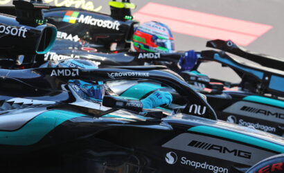 Close-up of Formula 1 cars lined up in a pit, with drivers in cockpits and visible sponsor logos like Microsoft, AMD, Petronas, and Snapdragon on sleek black-and-teal liveries.