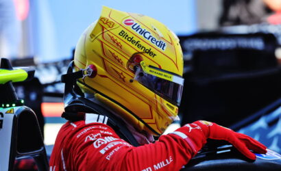 Close-up of a race car driver's yellow helmet with Unicredit and Bitdefender logos, visor down, red racing suit in a pit area.