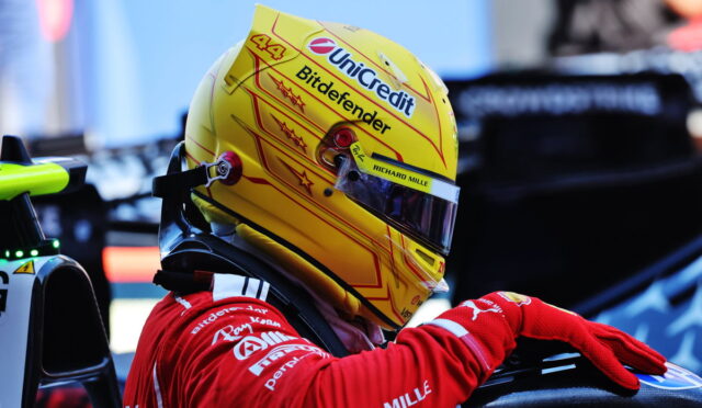 Close-up of a race car driver's yellow helmet with Unicredit and Bitdefender logos, visor down, red racing suit in a pit area.