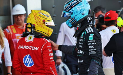 Two Formula 1 drivers face each other in the pit area; one in a red suit with 'HAMILTON' on the back and a yellow helmet, the other in a black Mercedes suit with a blue helmet.
