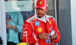 Ferrari Formula 1 driver in red racing suit and cap looking to the side, holding sunglasses in a pit area with sponsor logos visible on suit.