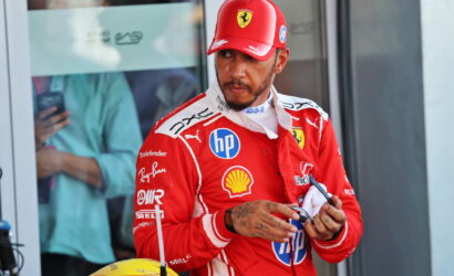 Ferrari Formula 1 driver in red racing suit and cap looking to the side, holding sunglasses in a pit area with sponsor logos visible on suit.