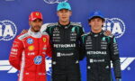 Three Formula 1 drivers posing on a podium: Ferrari driver in red on the left, and two Mercedes drivers in black Petronas suits in the center and right, against a blue sponsor backdrop, with team logos visible.