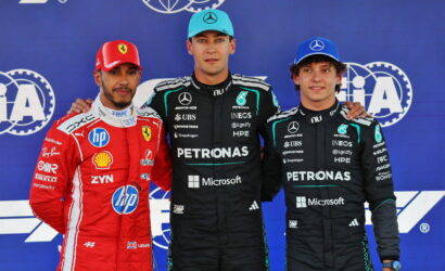 Three Formula 1 drivers posing on a podium: Ferrari driver in red on the left, and two Mercedes drivers in black Petronas suits in the center and right, against a blue sponsor backdrop, with team logos visible.