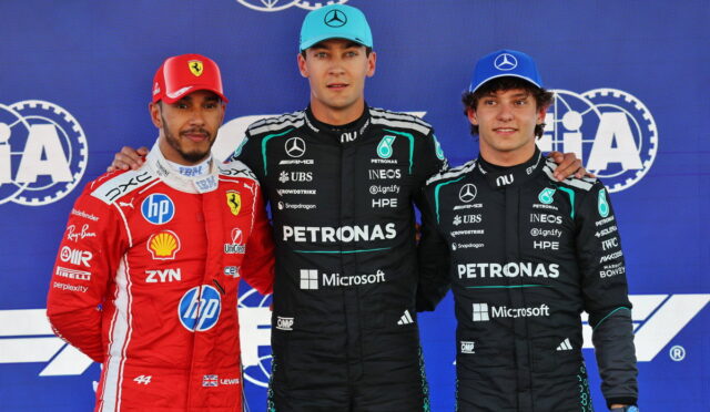 Three Formula 1 drivers posing on a podium: Ferrari driver in red on the left, and two Mercedes drivers in black Petronas suits in the center and right, against a blue sponsor backdrop, with team logos visible.