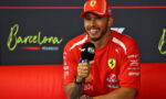 Ferrari driver in red interview shirt holding a microphone during a Barcelona event press conference, smiling at the camera atop a red seating backdrop.