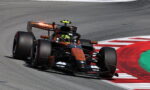 Formula 1 style race car in orange and black livery taking a sharp corner on a track with red‑and‑white curbs visible ahead, driver in helmet inside cockpit.