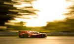 Red race car speeding along a track with motion blur in the background and guardrails on the side, during a golden-hour scene.