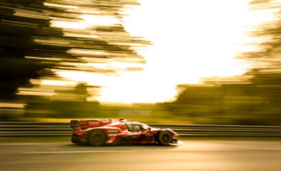 Red race car speeding along a track with motion blur in the background and guardrails on the side, during a golden-hour scene.