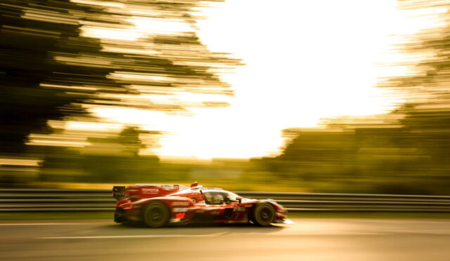 Red race car speeding along a track with motion blur in the background and guardrails on the side, during a golden-hour scene.