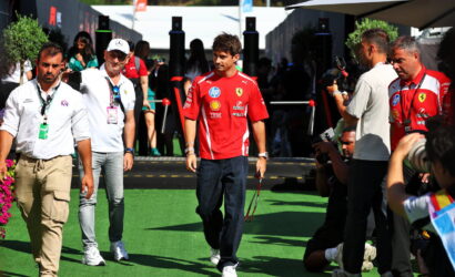 Ferrari team member in a red shirt walks on green turf as photographers capture him at a Formula 1 event by the paddock area.