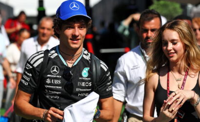 Young man wearing a black Mercedes racing jersey and blue cap walks through a crowd at an event, holding a white cloth.