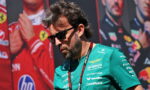 Man in teal Formula One team shirt and sunglasses, looking down with a lanyard, race posters in the background at a motorsport event.