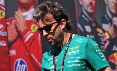 Man in teal Formula One team shirt and sunglasses, looking down with a lanyard, race posters in the background at a motorsport event.