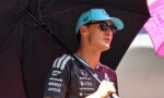 Man in a branded racing jersey and blue cap stands under a large black umbrella, shielding from the sun at an outdoor motorsport event.