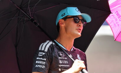 Man in a branded racing jersey and blue cap stands under a large black umbrella, shielding from the sun at an outdoor motorsport event.
