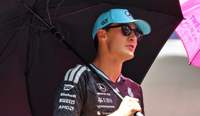 Man in a branded racing jersey and blue cap stands under a large black umbrella, shielding from the sun at an outdoor motorsport event.