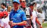 Male race team member in a blue cap and polo, holding a water bottle in a crowded pit area with spectators behind him.