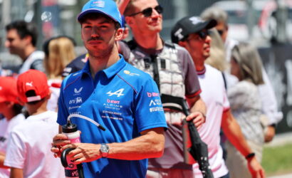 Male race team member in a blue cap and polo, holding a water bottle in a crowded pit area with spectators behind him.