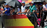 Racers and team members stand behind a barrier with a dotted German flag graphic at a race track pit area, holding umbrellas and chatting.