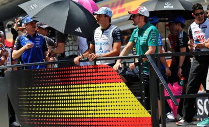 Racers and team members stand behind a barrier with a dotted German flag graphic at a race track pit area, holding umbrellas and chatting.