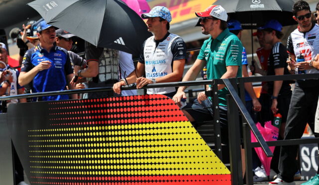 Racers and team members stand behind a barrier with a dotted German flag graphic at a race track pit area, holding umbrellas and chatting.